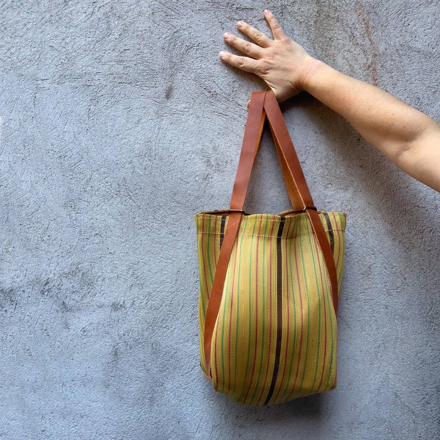 Picture of yellow striped tote bag against a textured blue background.with a hand and forearm holding the leather straps.