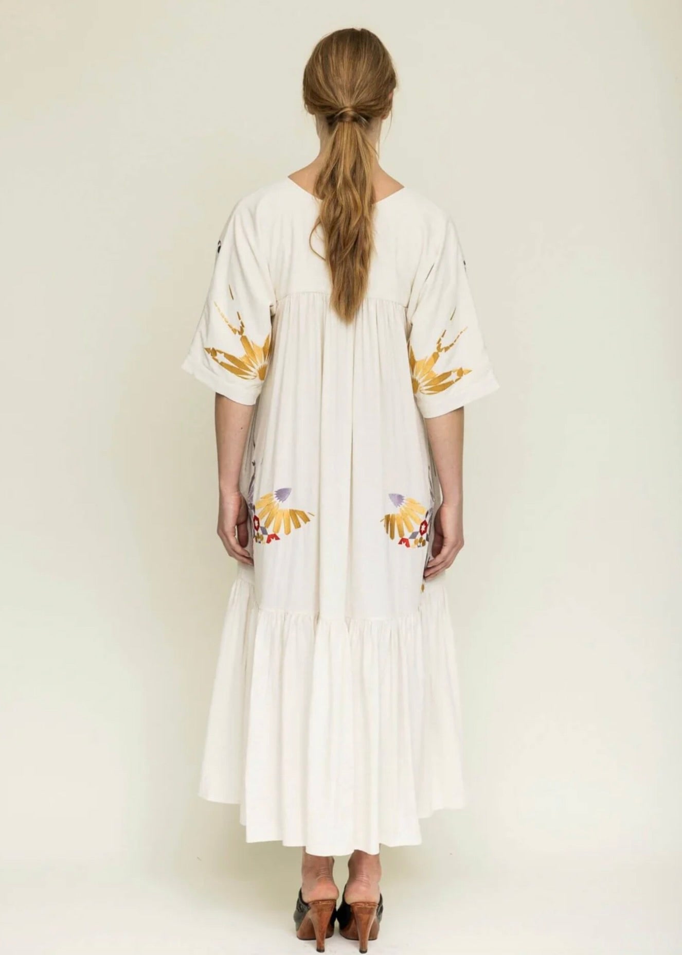 View from behind of woman with blond hair tied back wearing a midi length cream colored dress with colorful embroidery around the yolk, sleeve and hips.
