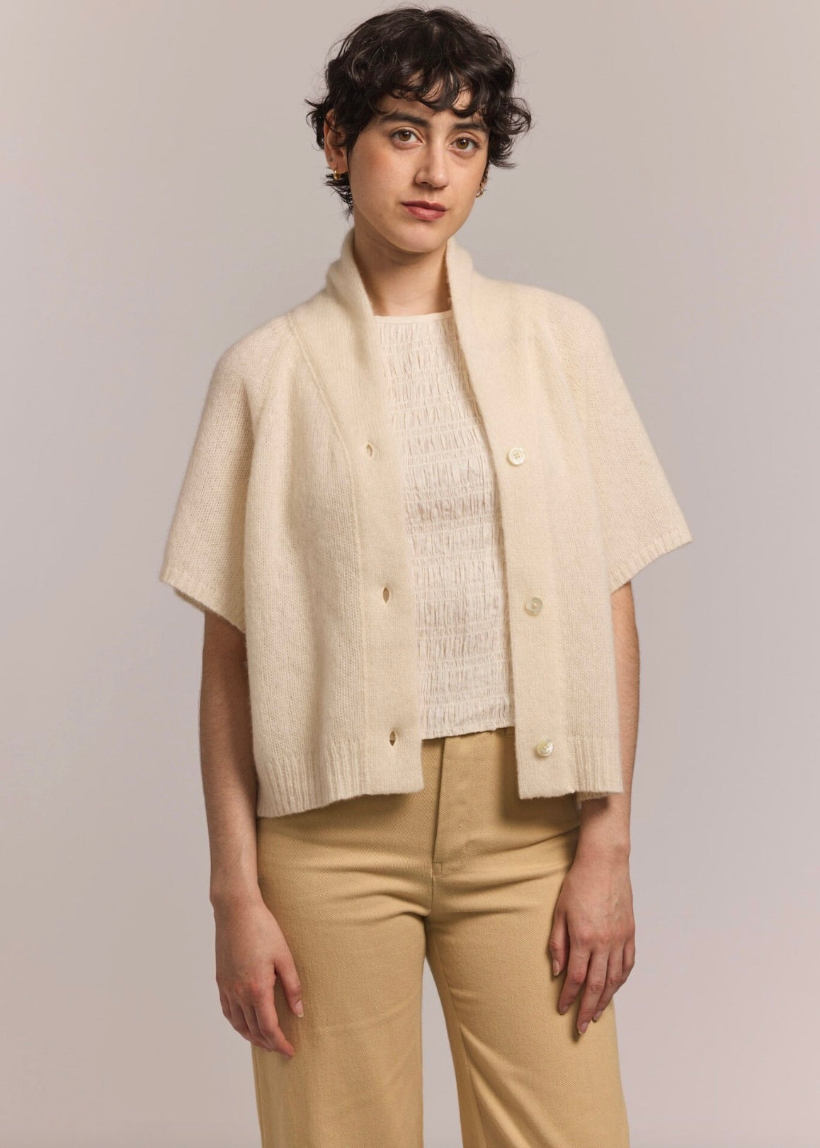 Woman with short wavy dark brown hair wearing an ivory short sleeve cashmere cardigan with a silk blouse and tan colored pants