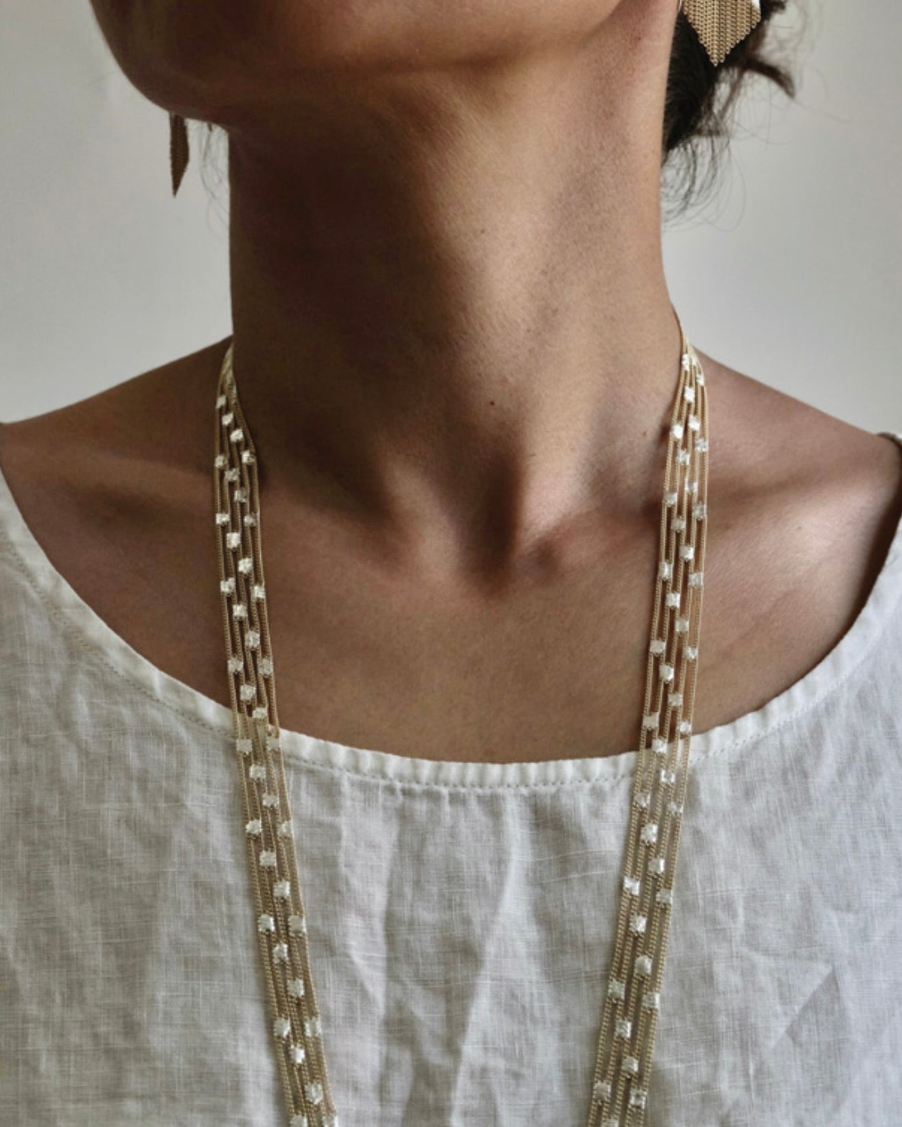 Close detail of cropped image of woman with long dark hair wearing a white round neck shirt and, silver earrings and a long silver and brass chain necklace.