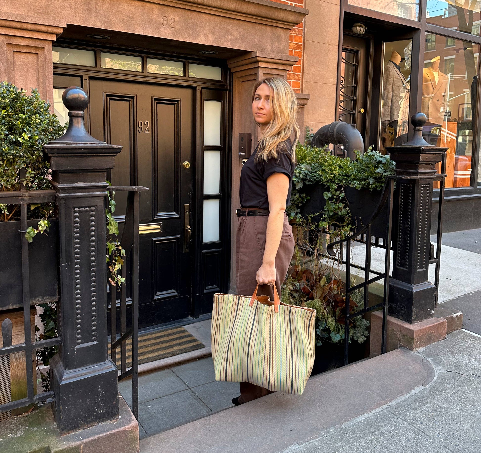 Picture of young women with blond hair and blue eyes holding a striped tote bag at the entrance stairs to a reb brick townhouse.