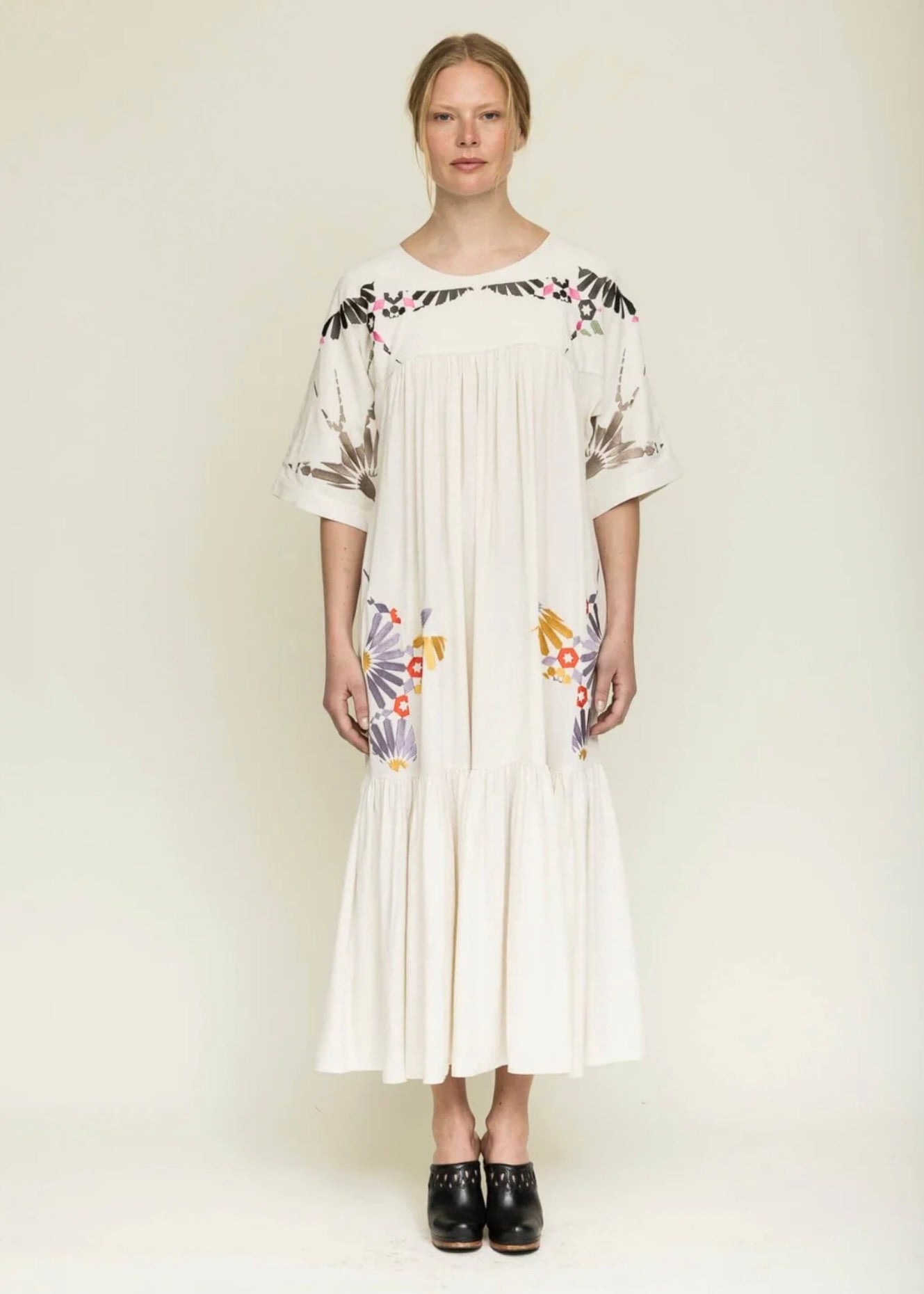woman with blond hair tied back wearing a midi length cream colored dress with colorful embroidery around the yolk and hips.