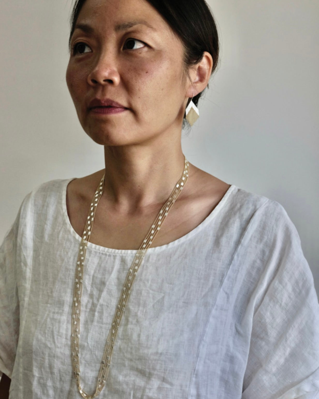 Cropped image of woman with long dark hair wearing a white round neck shirt and, silver earrings and a long silver and brass chain necklace.