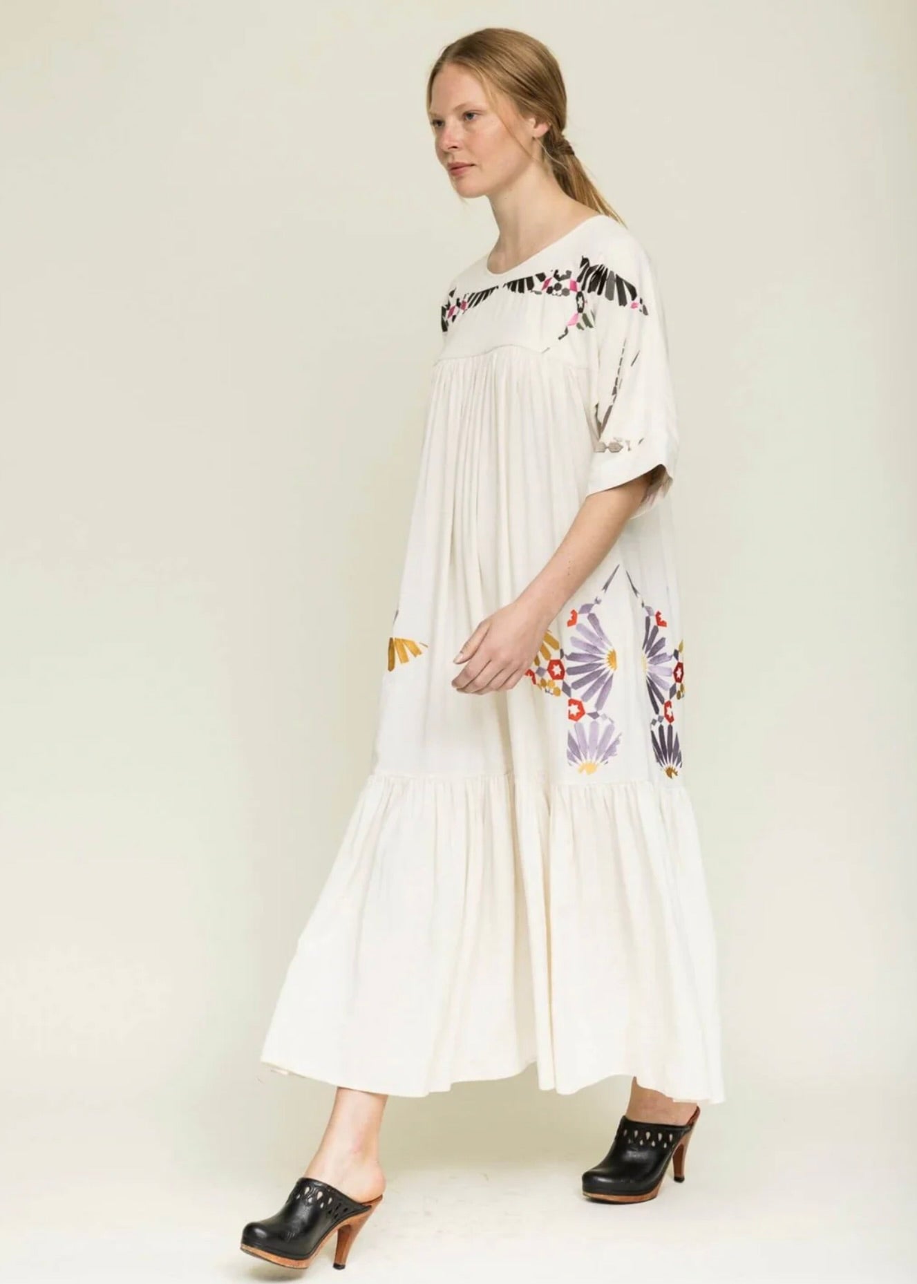 woman stepping forward with blond hair tied back wearing a midi length cream colored dress with colorful embroidery around the yolk and hips.