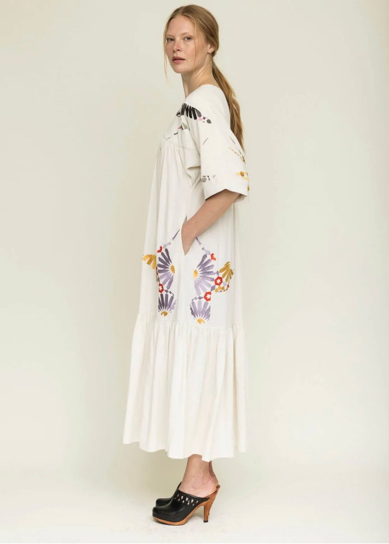 Side view if woman with blond hair tied back wearing a midi length cream colored dress with colorful embroidery around the yolk and hips.