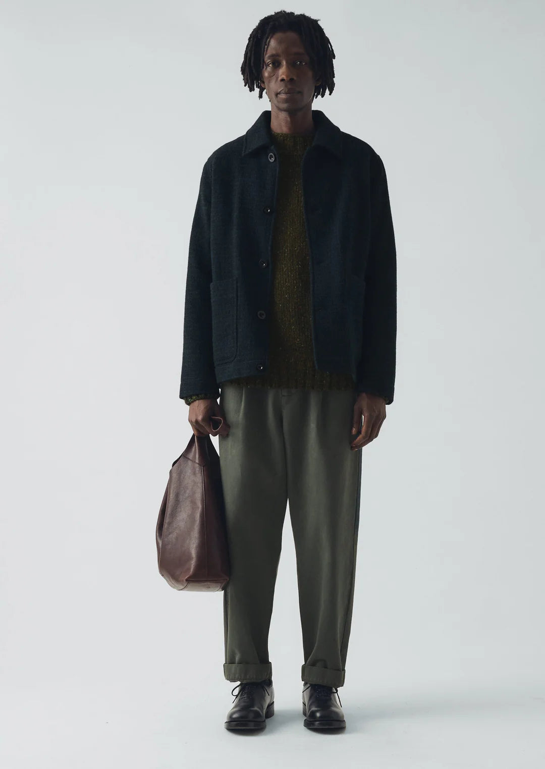 Man holding a brown leather bag wearing a dark jacket and green pants on a white background