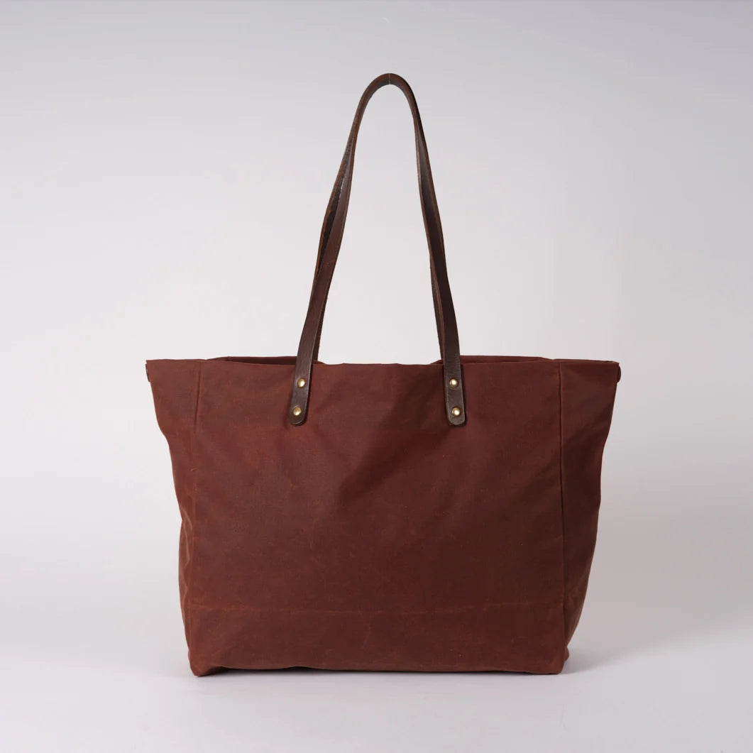 A brown canvas tote bag with dark brown leather shoulder straps and silver-tone metal rivets, photographed against a neutral light gray background.