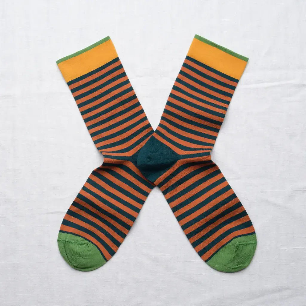 A pair of striped crew socks arranged in an X shape on a white fabric background. The socks feature horizontal stripes of dark teal and orange-brown, with a solid dark teal center, green toes, and yellow-green cuffs.