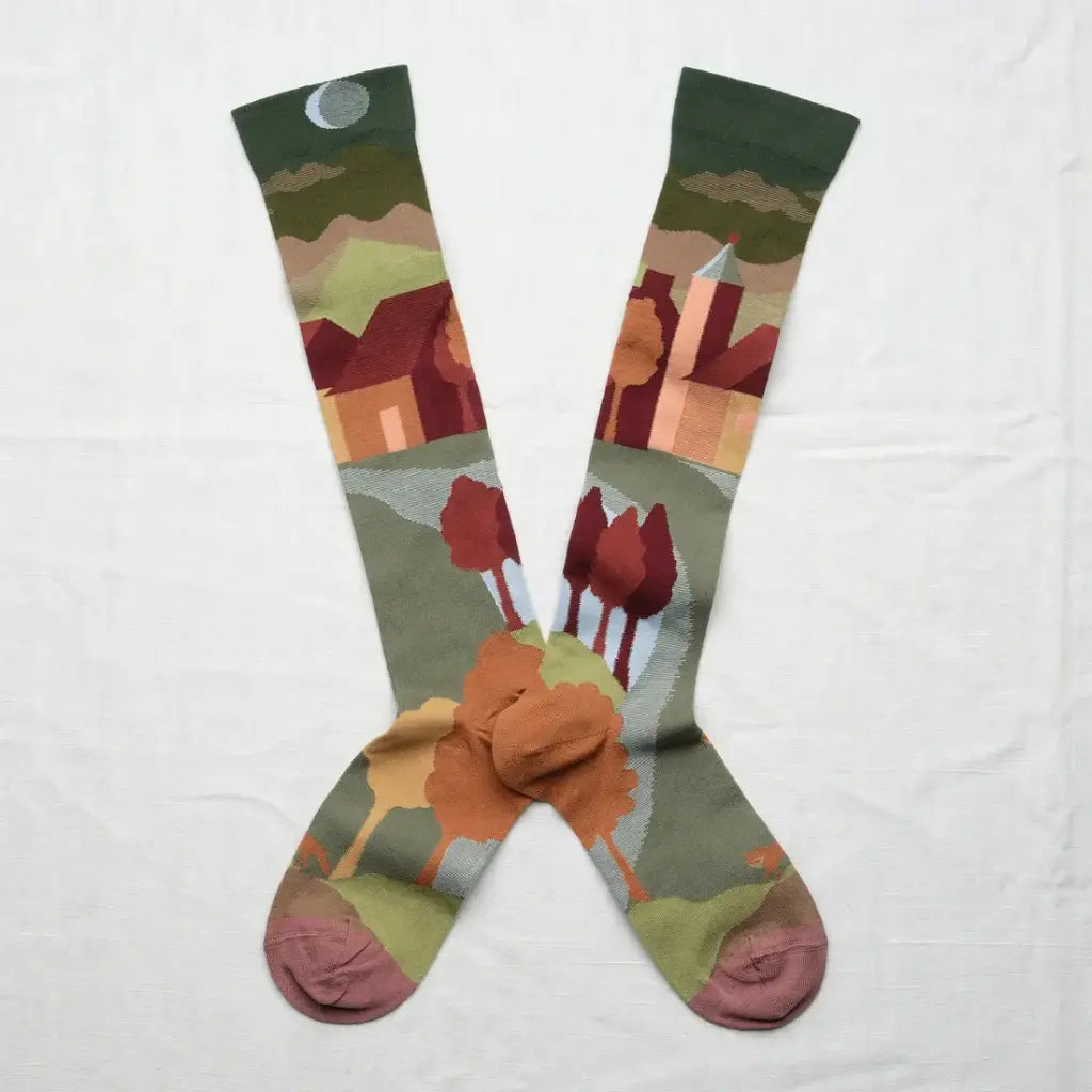 A pair of long crew socks laid flat in an X shape on a white fabric background, featuring a stylized landscape print with trees, houses, mountains, and a moon.