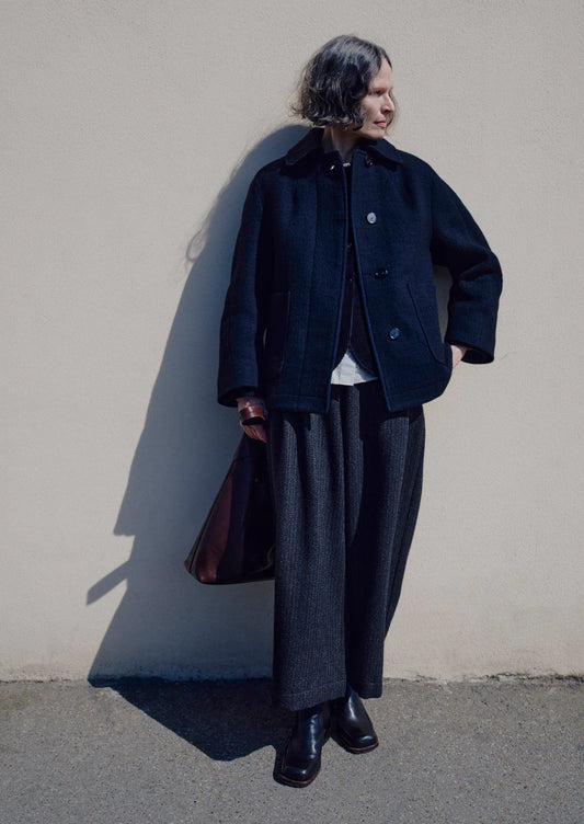 Person wearing a dark coat and gray pants against a plain wall.