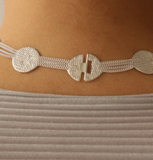 A close-up of a textured silver choker necklace worn on a person's neck, featuring circular disc elements and a central clasp, above a ribbed light gray top.