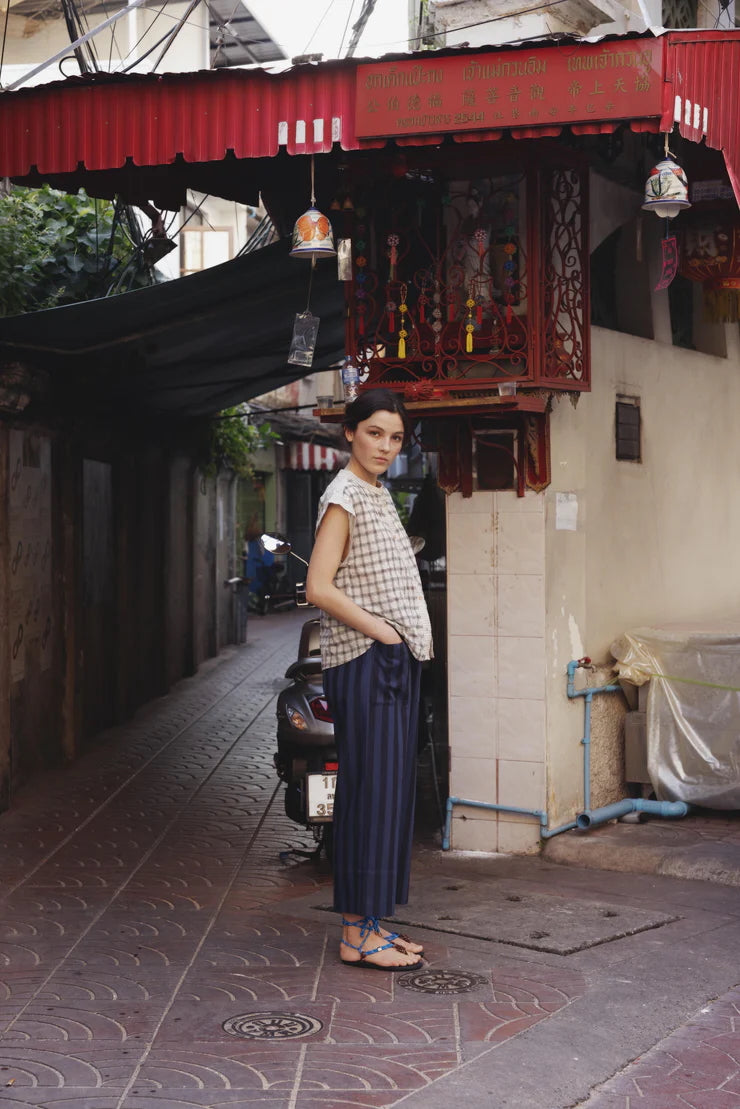 Picture of young woman with short black hair and blue eyes standing in front of a oriental alley with tiled road and a red covered roof. Wearing a a white checked sleeveless shirt and cream pants.
