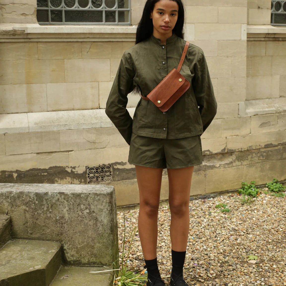 Woman wearing green shorts and jacket with small crossbody handmade brown leather bag with two brass pop closures.