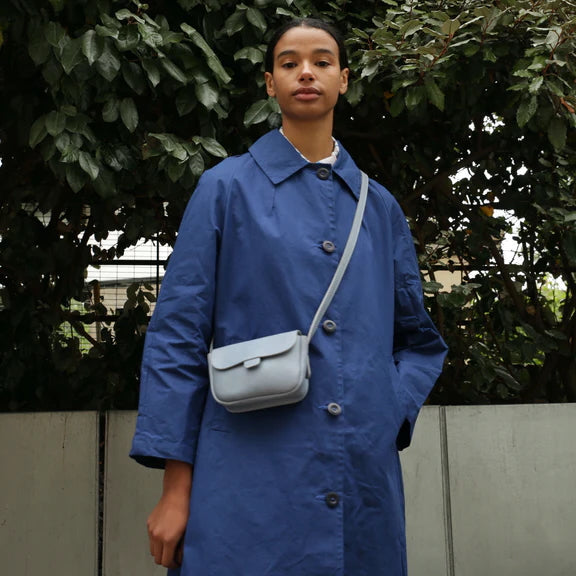 Picture of cornflower blue handbag front view. Being worn by a women with short black hair and brown eyes. Wearing a blue Macintosh.