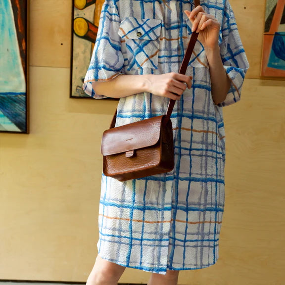 Picture, mid shoot of the front of a brown leather handbag being worn by a person in a checked blue dress.