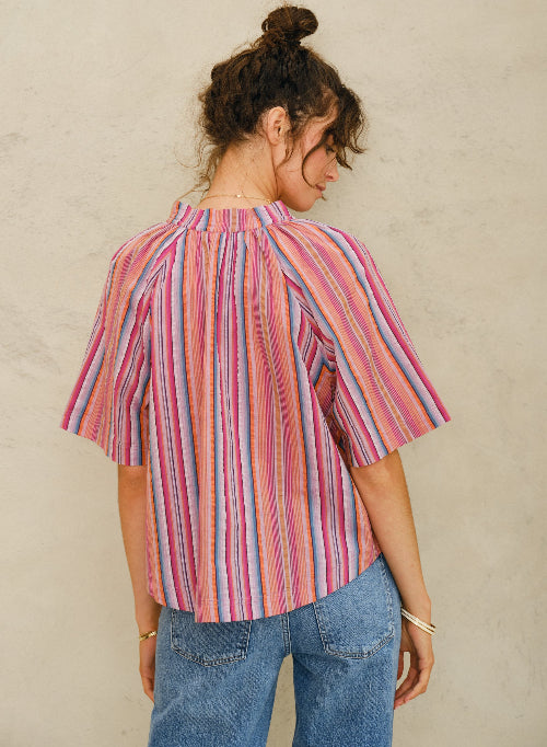 Picture of woman, from behind. With curly brown hair tied up in a bun and brown eyes. Wearing a stripy short sleeve cotton top and blue denim jeans. Standing against a stone colored background.