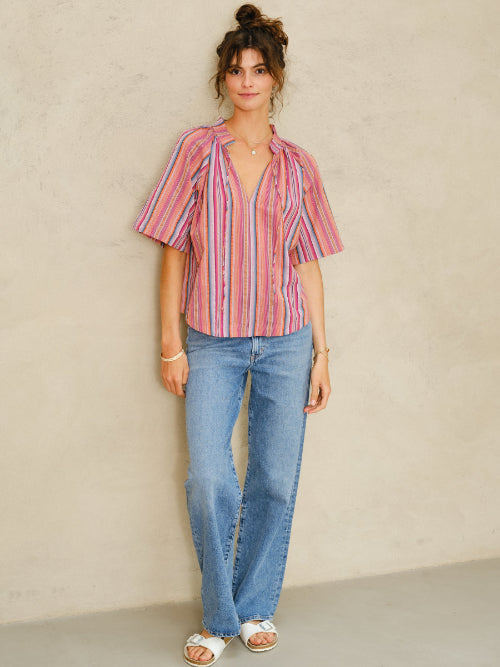 Picture of woman, full length. With curly brown hair tied up in a bun and brown eyes. Wearing a stripy short sleeve cotton top and blue denim jeans. Standing against a stone colored background.