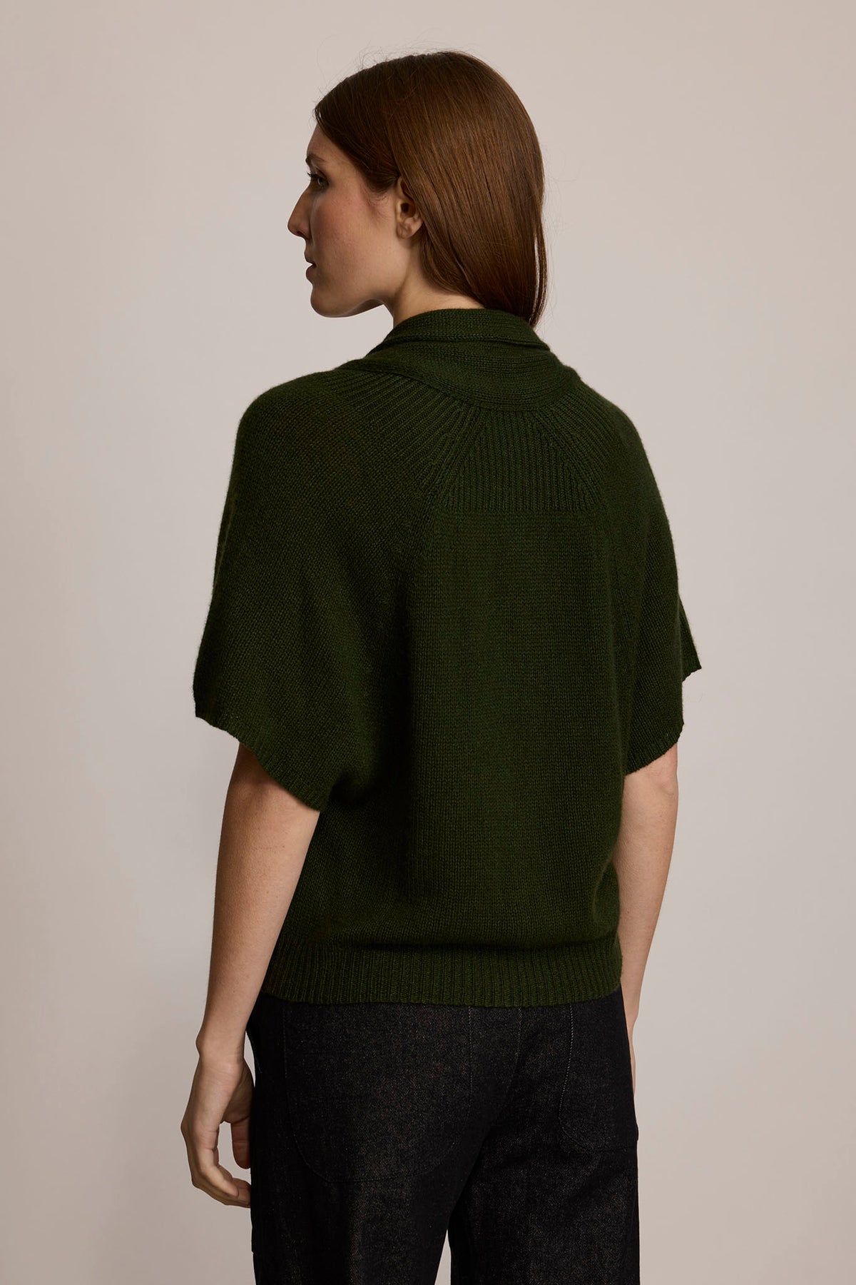 A view of the back of a model wearing a forest green cardigan and black denim pants.