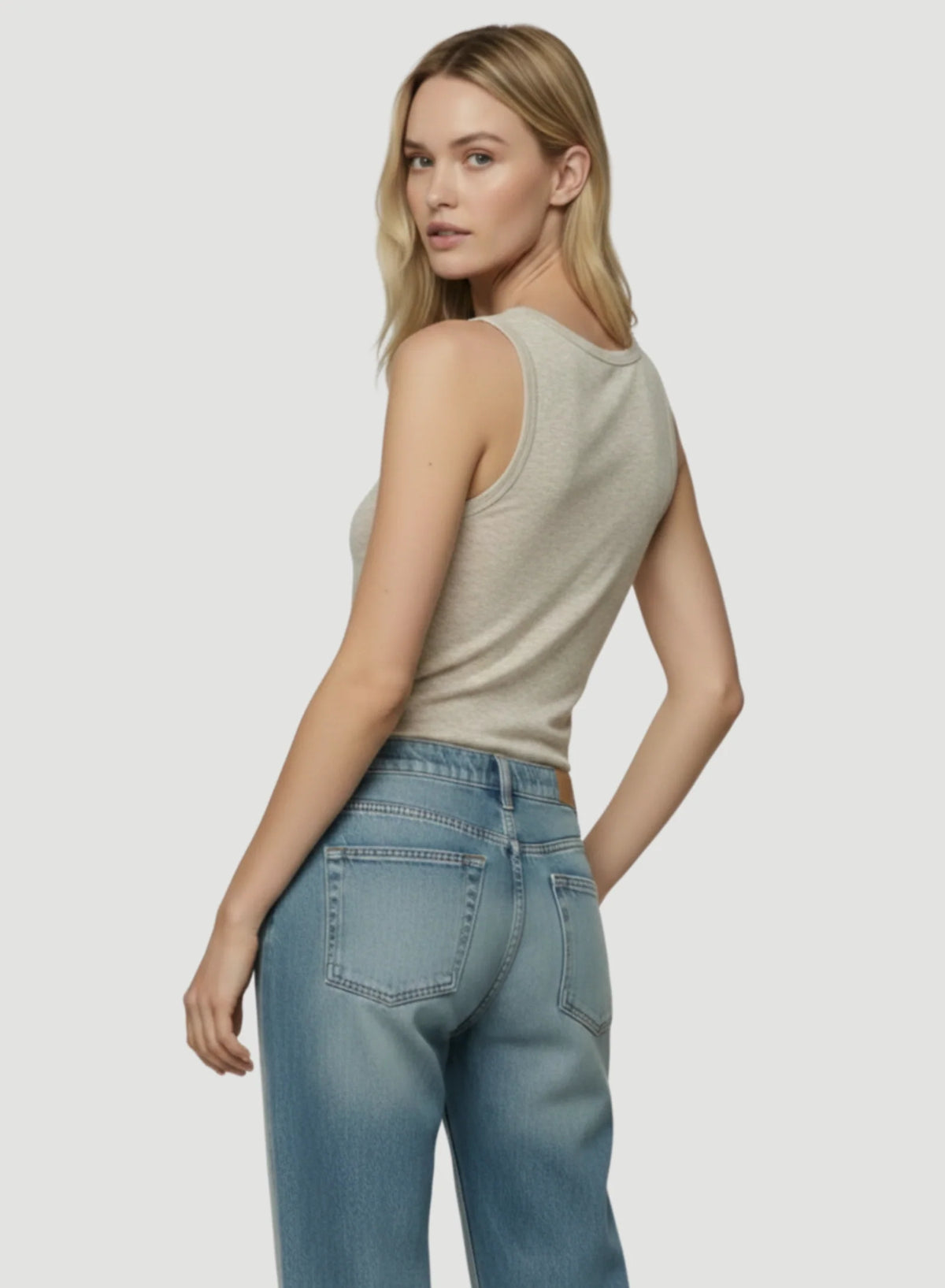 A model poses against a white background wearing a light beige tank top with wide straps and blue jeans.