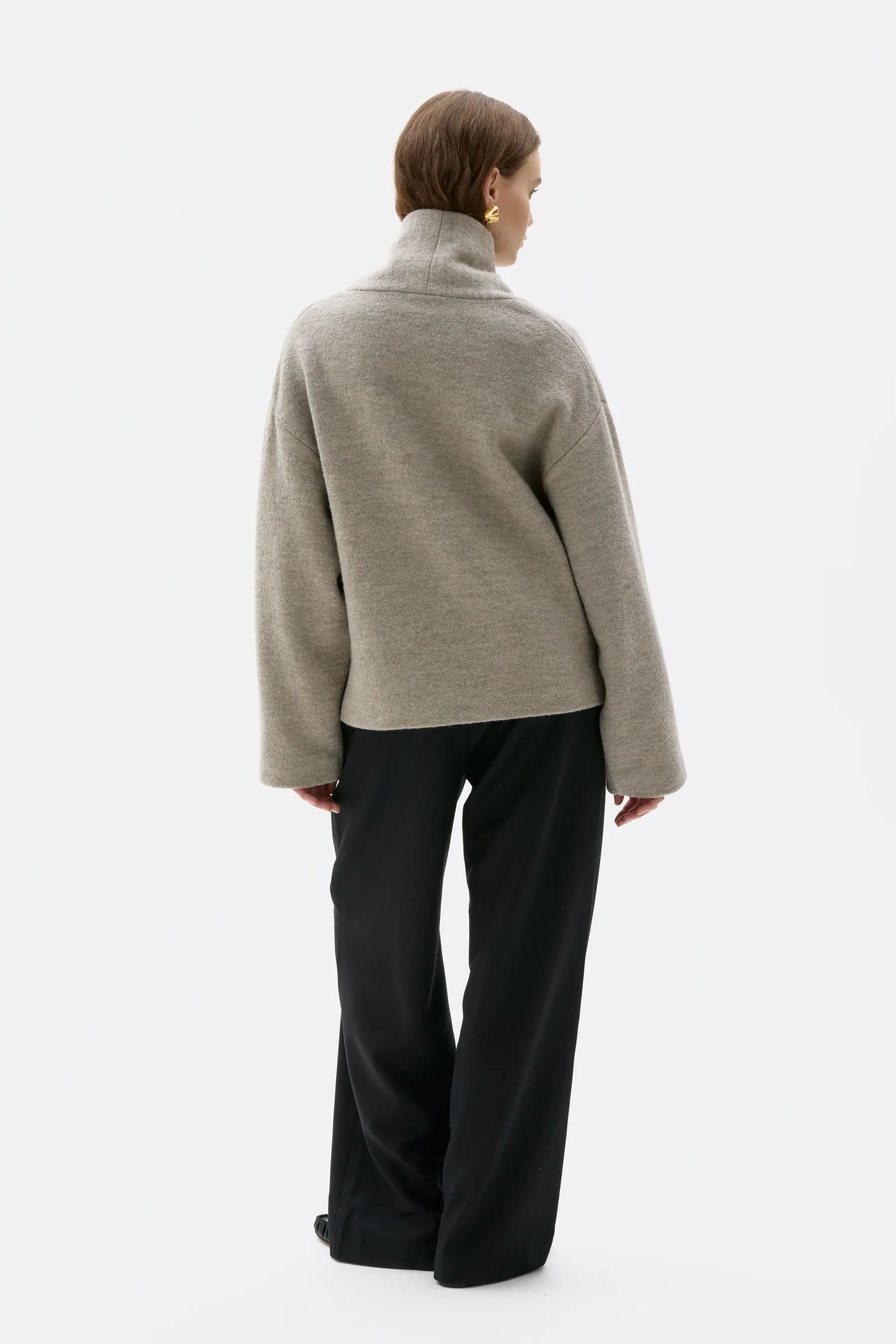 A model shown from the back wearing a light gray turtleneck sweater and black wide-leg trousers against a plain white studio background.
