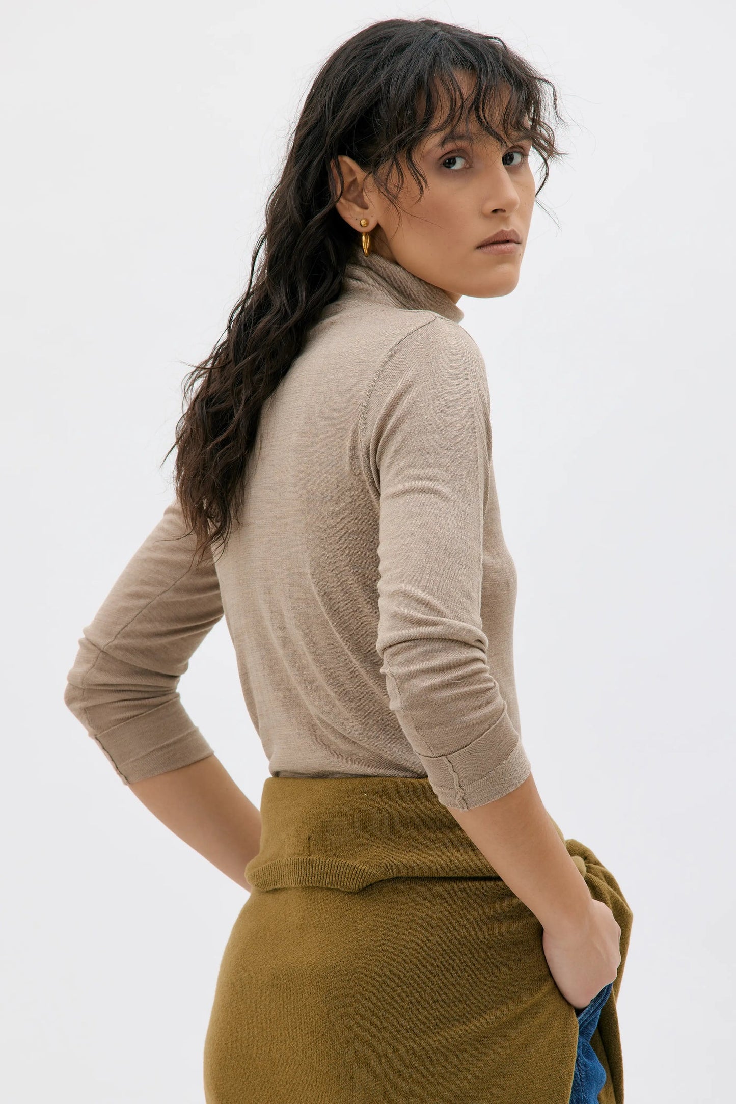 A woman wearing a beige ribbed turtleneck sweater and an olive green skirt, photographed from the back against a clean white studio background.