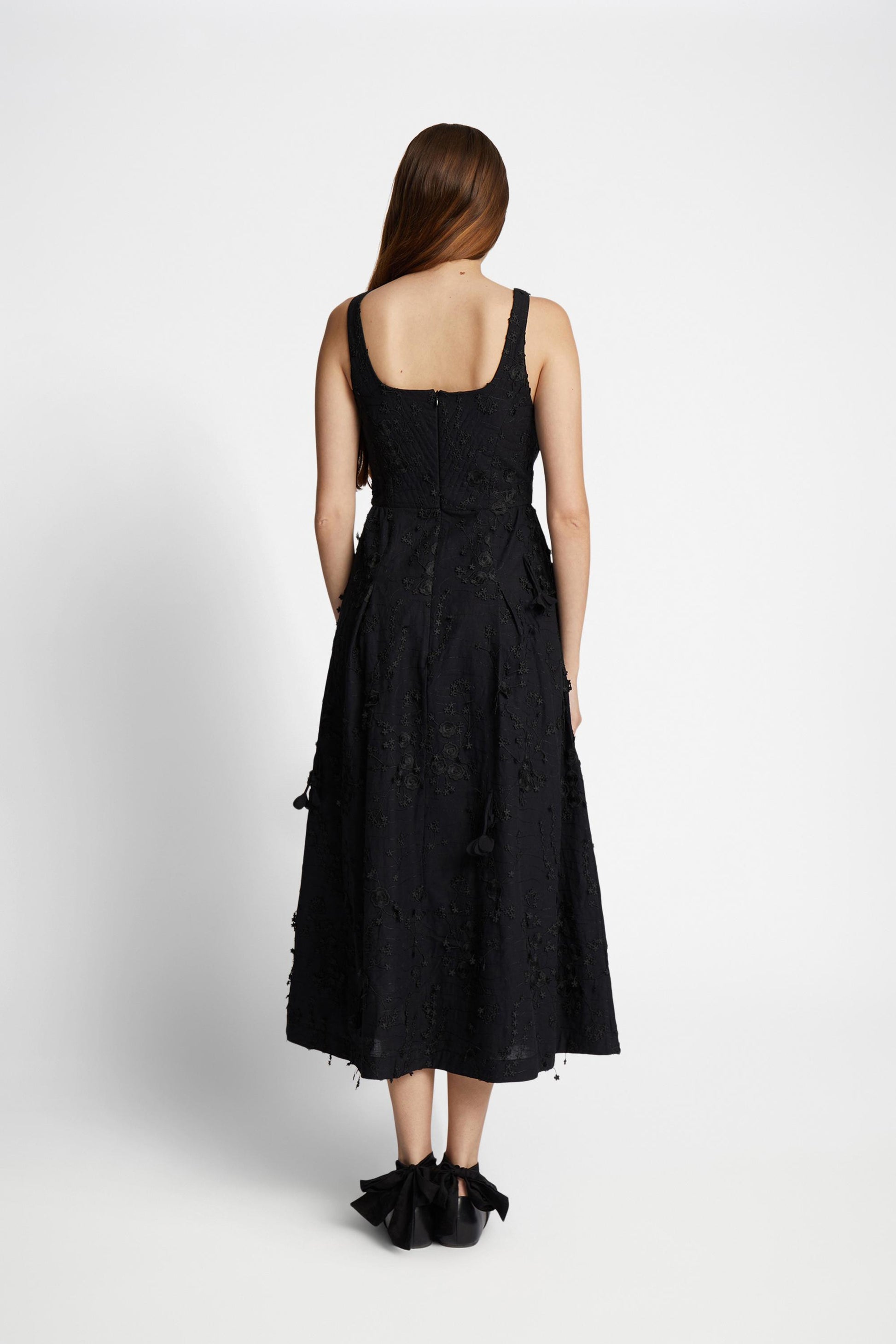 Reverse view of a woman wearing ablack dress with narrow straps embellished with floral applique all over.