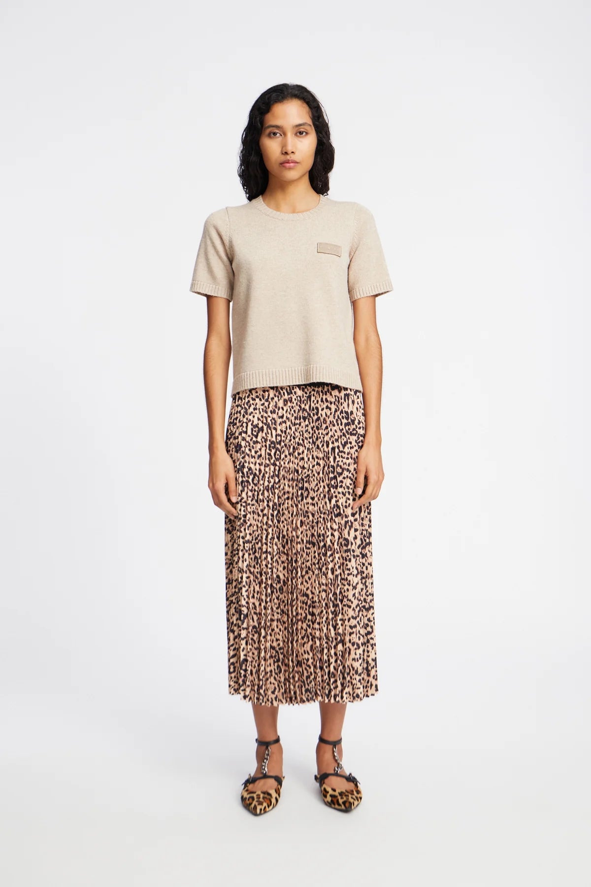 Picture of young women standing facing forward against a white background with dark brown eyes and black shoulder length hair. Wearing a leopard print pleated skirt and a light brown short sleeve knit.