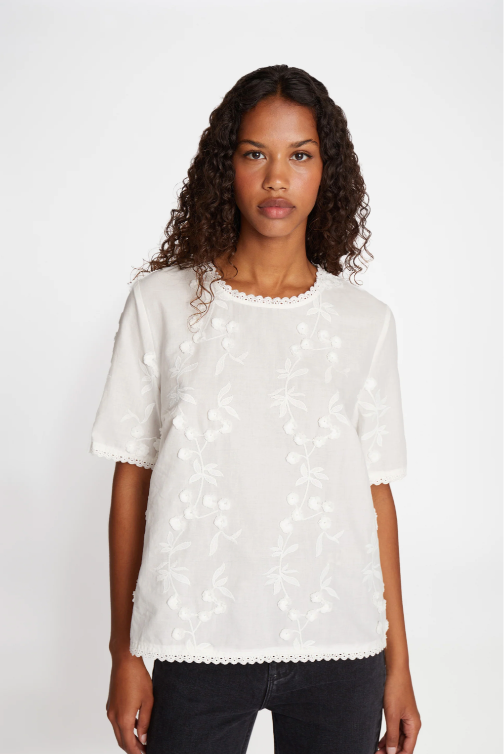 Woman with long dark wavy hair wearing white round neck blouse with floral decoration.