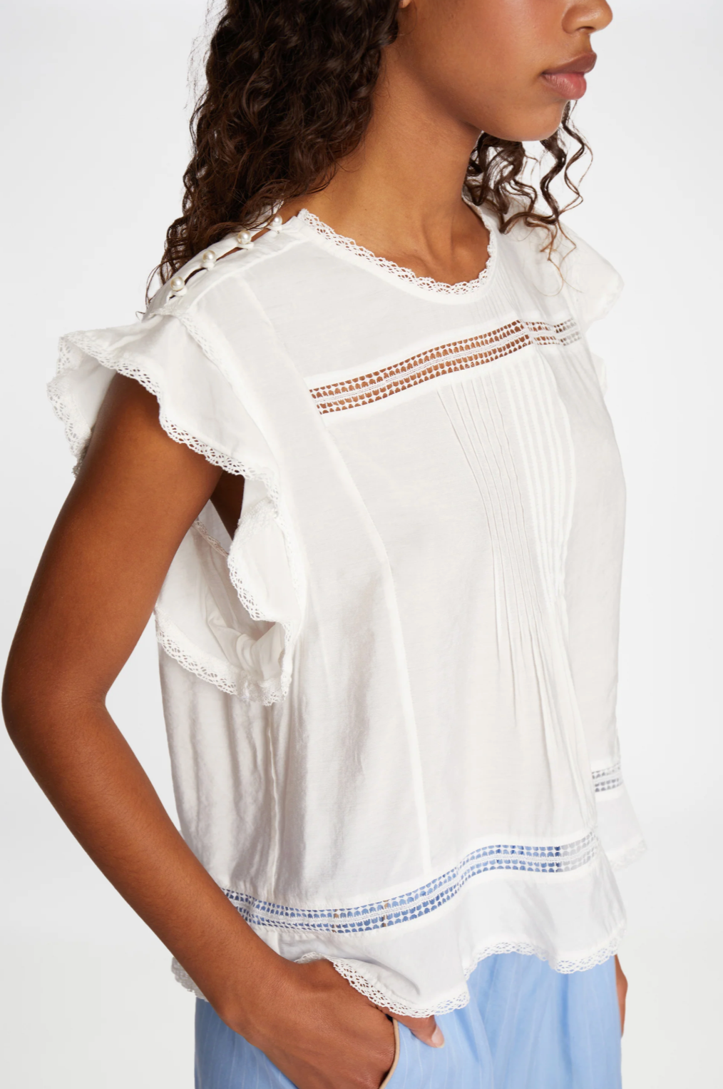 Cropped image of woman with long brown way hair wearing a white top short sleeve top with frills on capped sleeve and pearl buttons across one shoulder