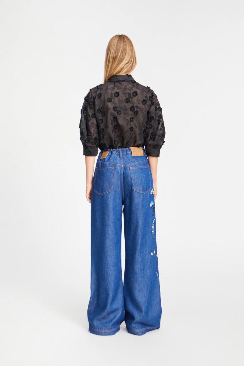 Picture of young women standing facing away, with fair skin and long shoulder length straight blond hair. wearing a black short sleeved shirt with black embroidered flowers and blue denim pants.