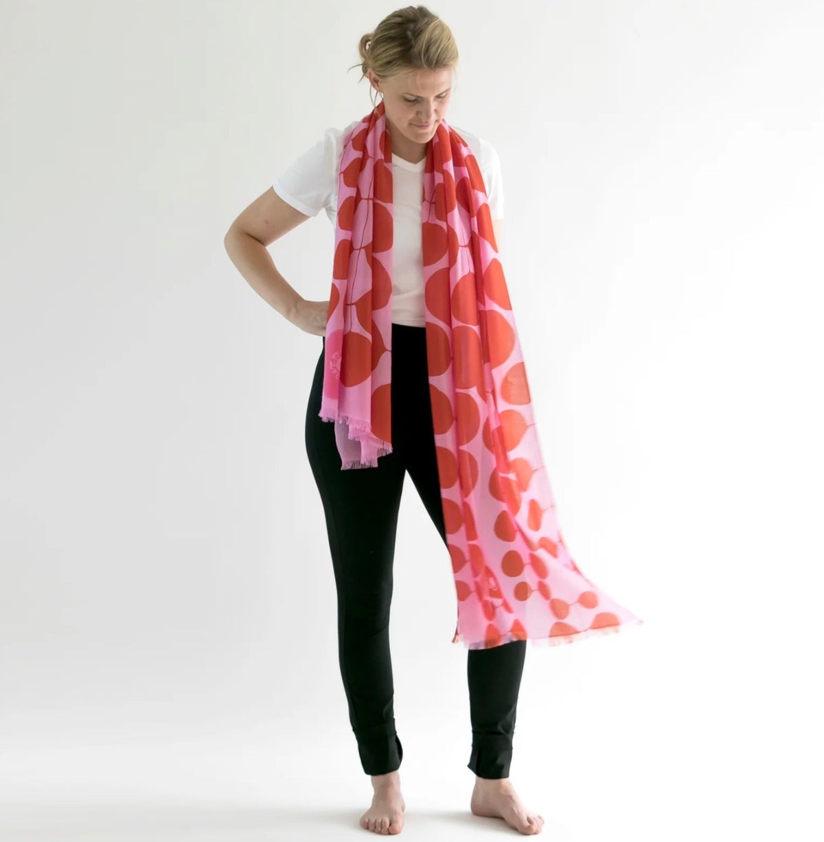 A long pink and red scarf shown in a playful pattern and wrapped around a woman with blonde hair and black pants.