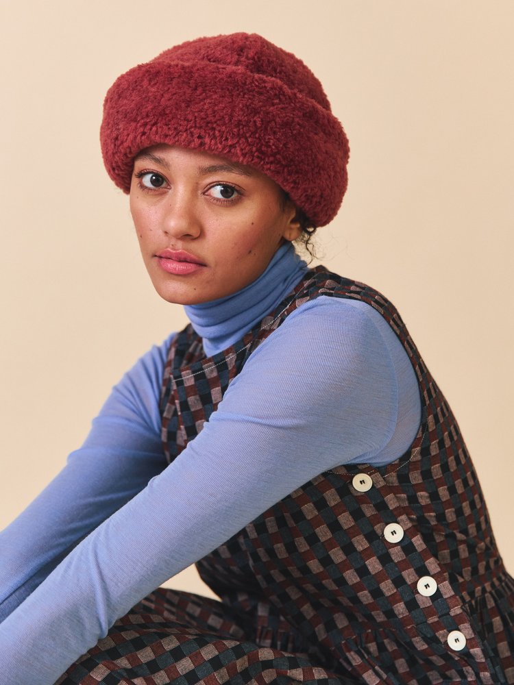 Person wearing a red knit hat, blue sweater, and patterned overalls against a beige background