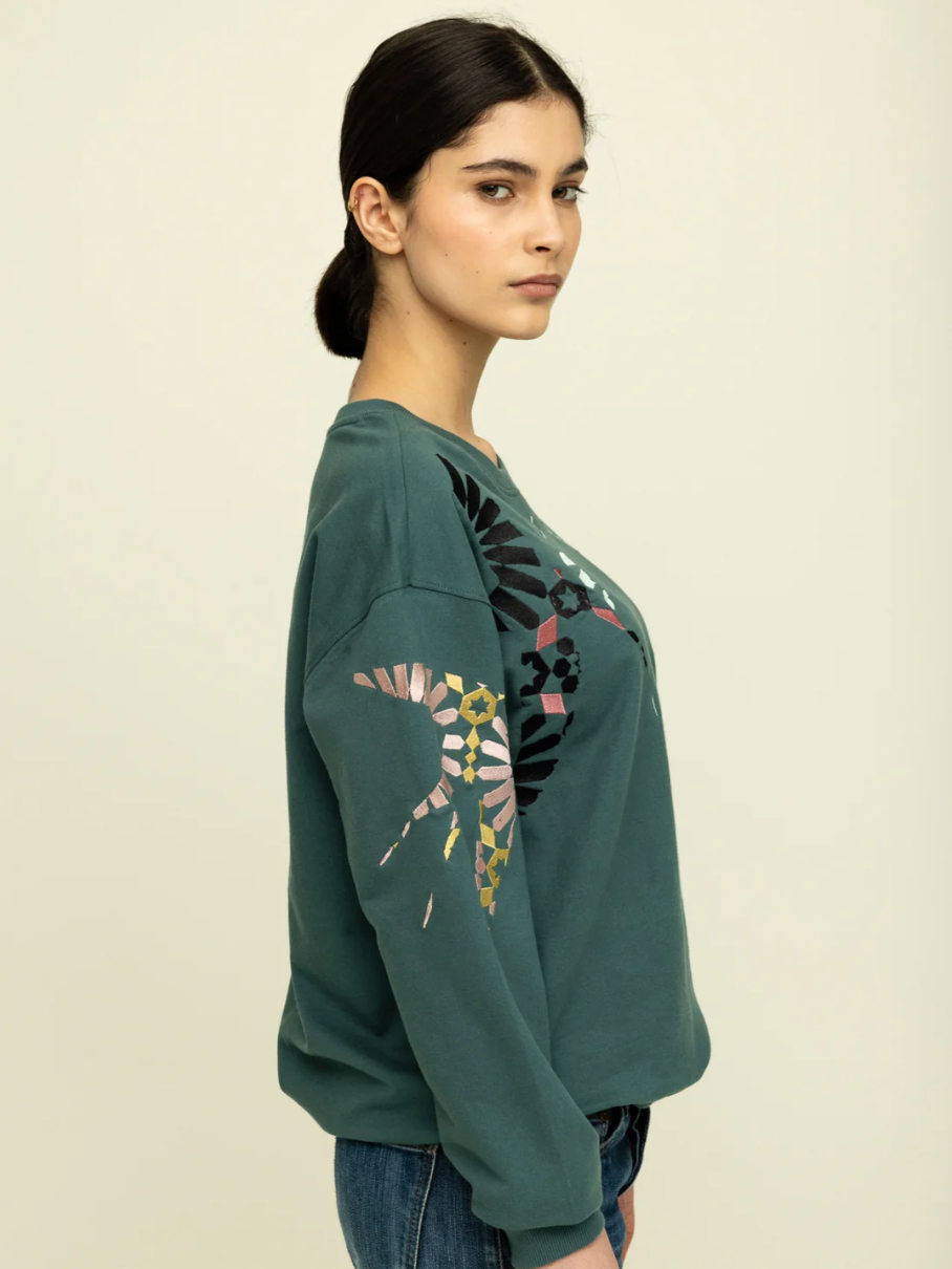 side view of cropped image of woman with brown hair tied back wearing dark green sweatshirt with pink, yellow, black and light green embroidery