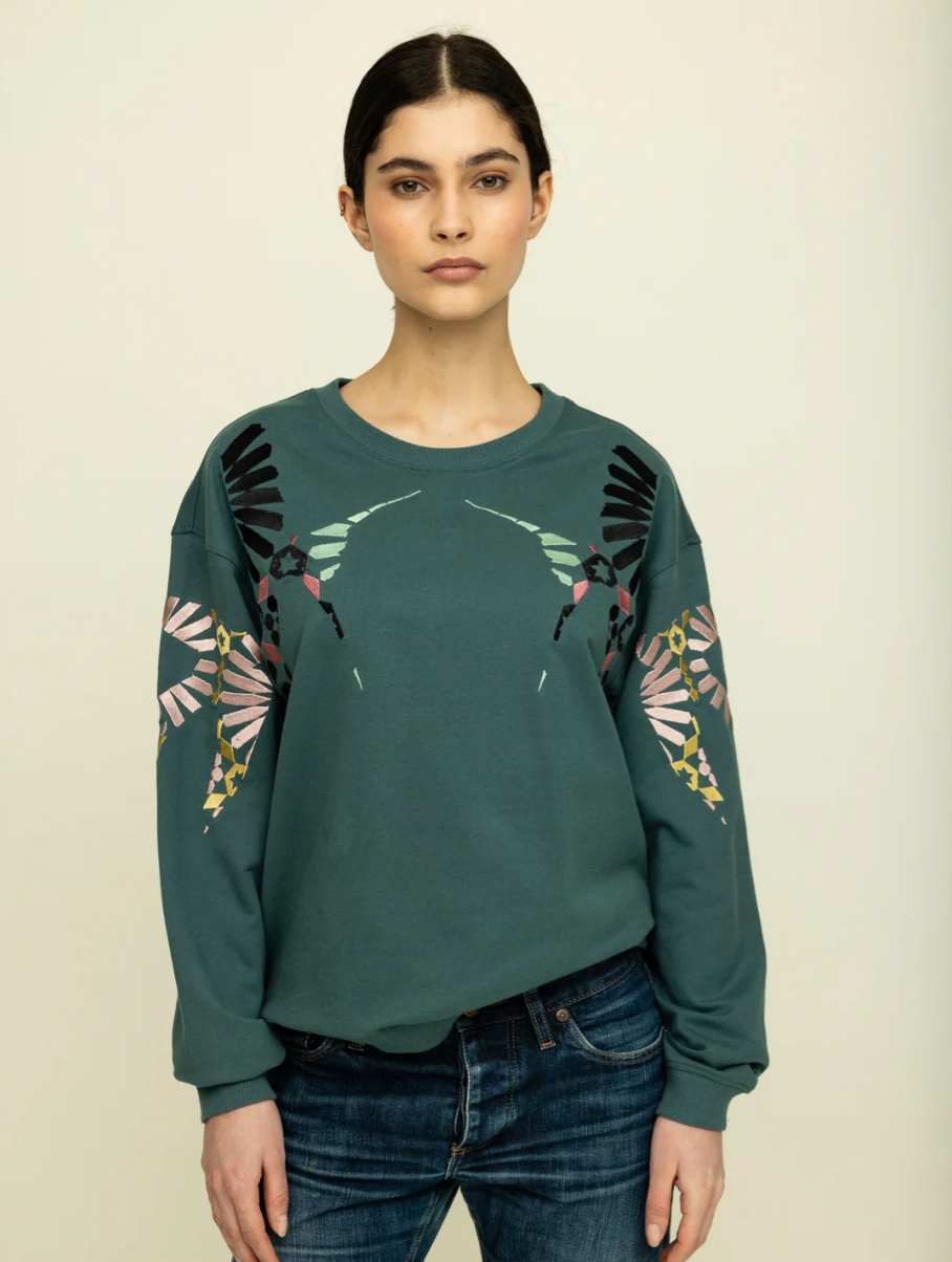 cropped image of woman with brown hair tied back wearing dark green sweatshirt with pink, yellow, black and light green embroidery