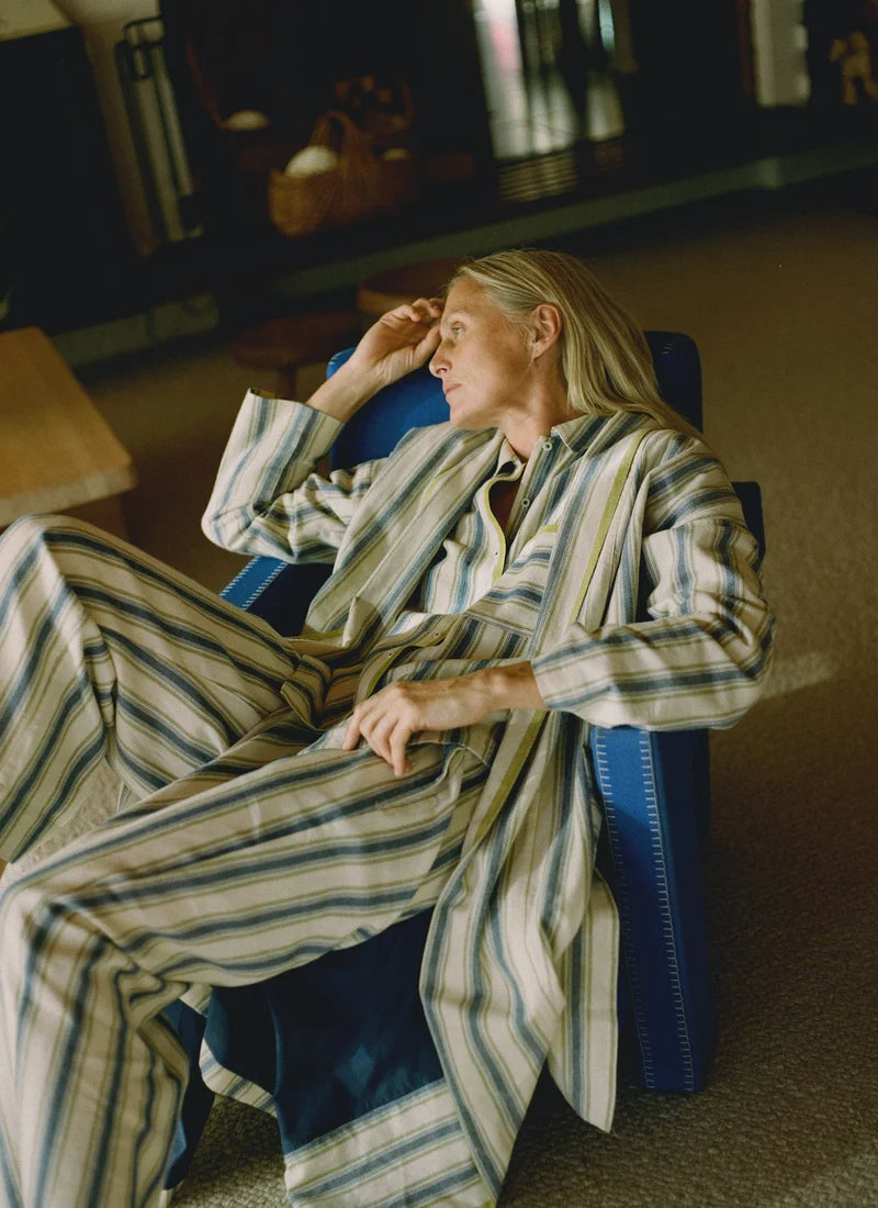 A model sitting in a blue chair and wearing a pajama set with blue and cream vertical stripes and a gooseberry-colored trim.