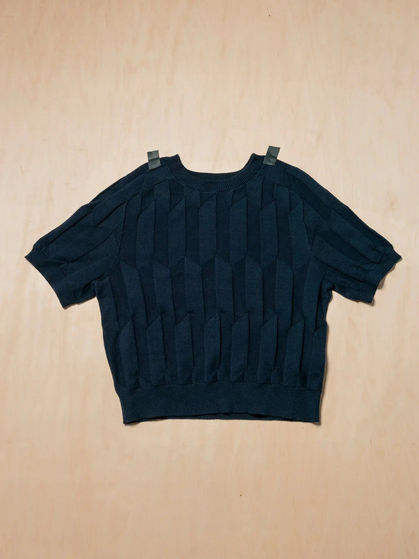 A dark navy blue short-sleeve knit top with a textured vertical pleated pattern, laid flat on a light wood background.