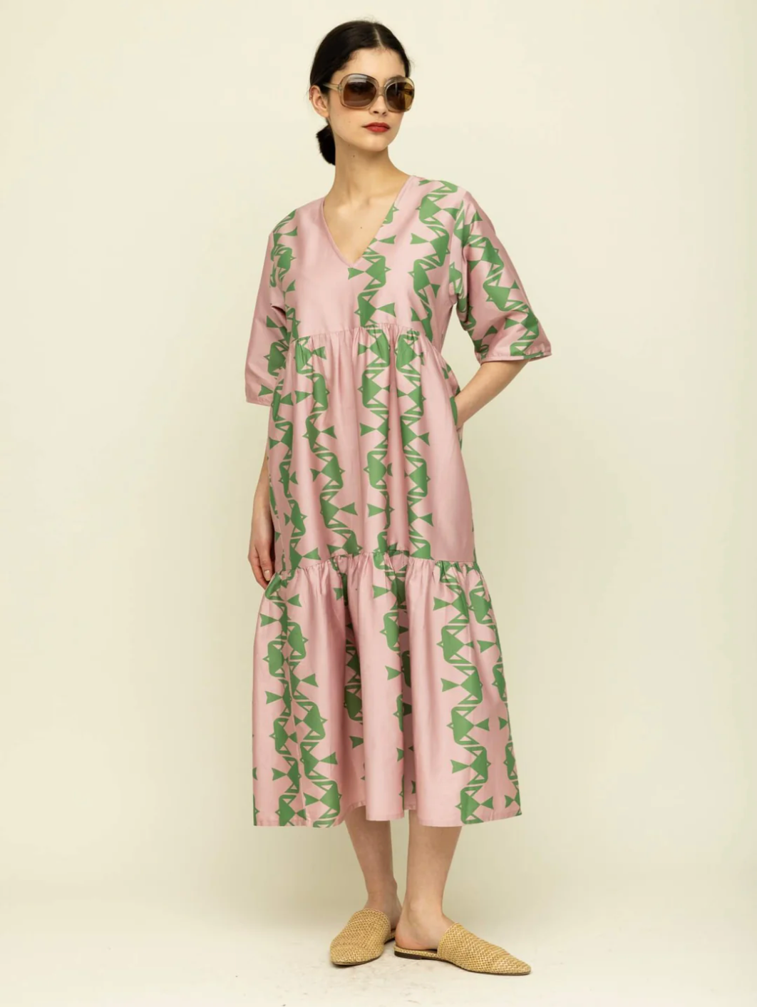 woman with shoulder length dark hair pulled back from her face wearing a midi length pink tiered dress with green abstract screen print design. She is wearing woven leather flat slipper sandals.