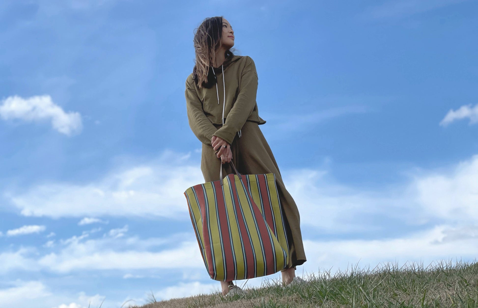 Picture of young women in a field  with blue cloudy sky holding purple tote bag on a white background.