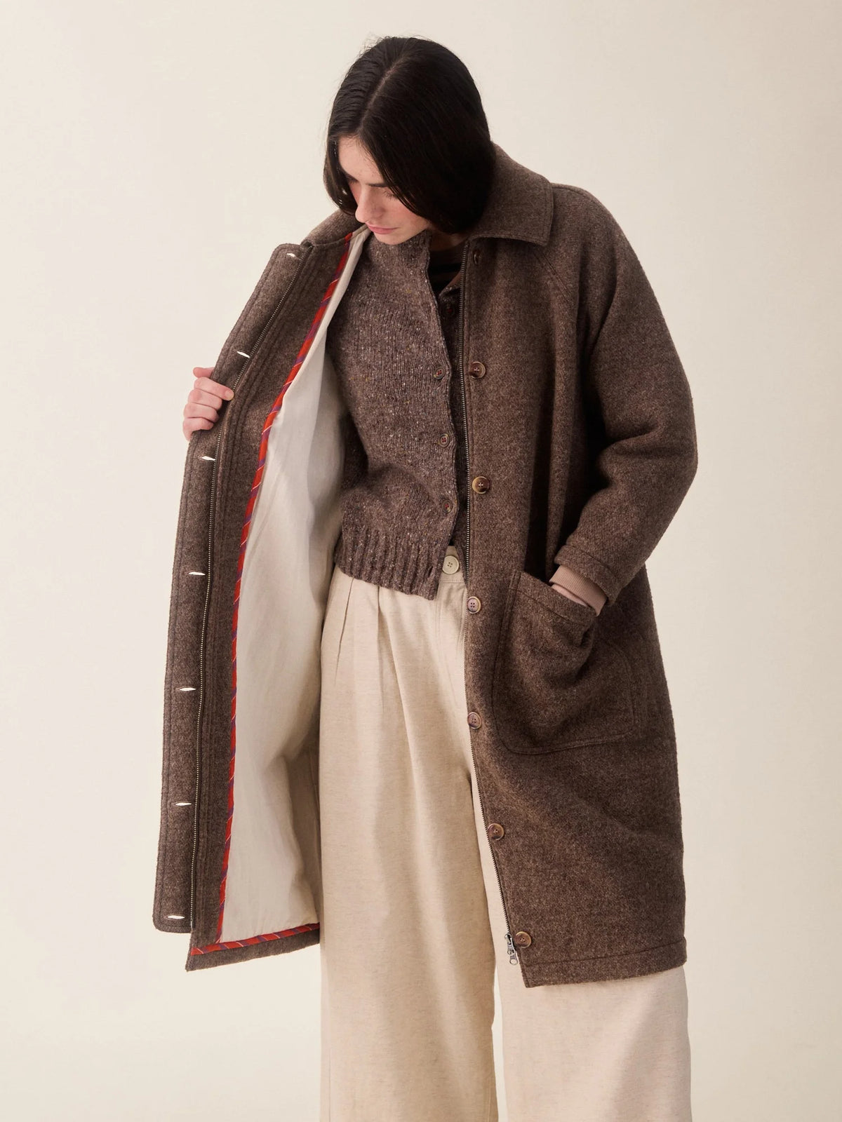 A model wearing a brown wool-blend button-front cardigan over a matching brown sweater and cream wide-leg trousers, holding open the cardigan to show the cream lining with red piping.