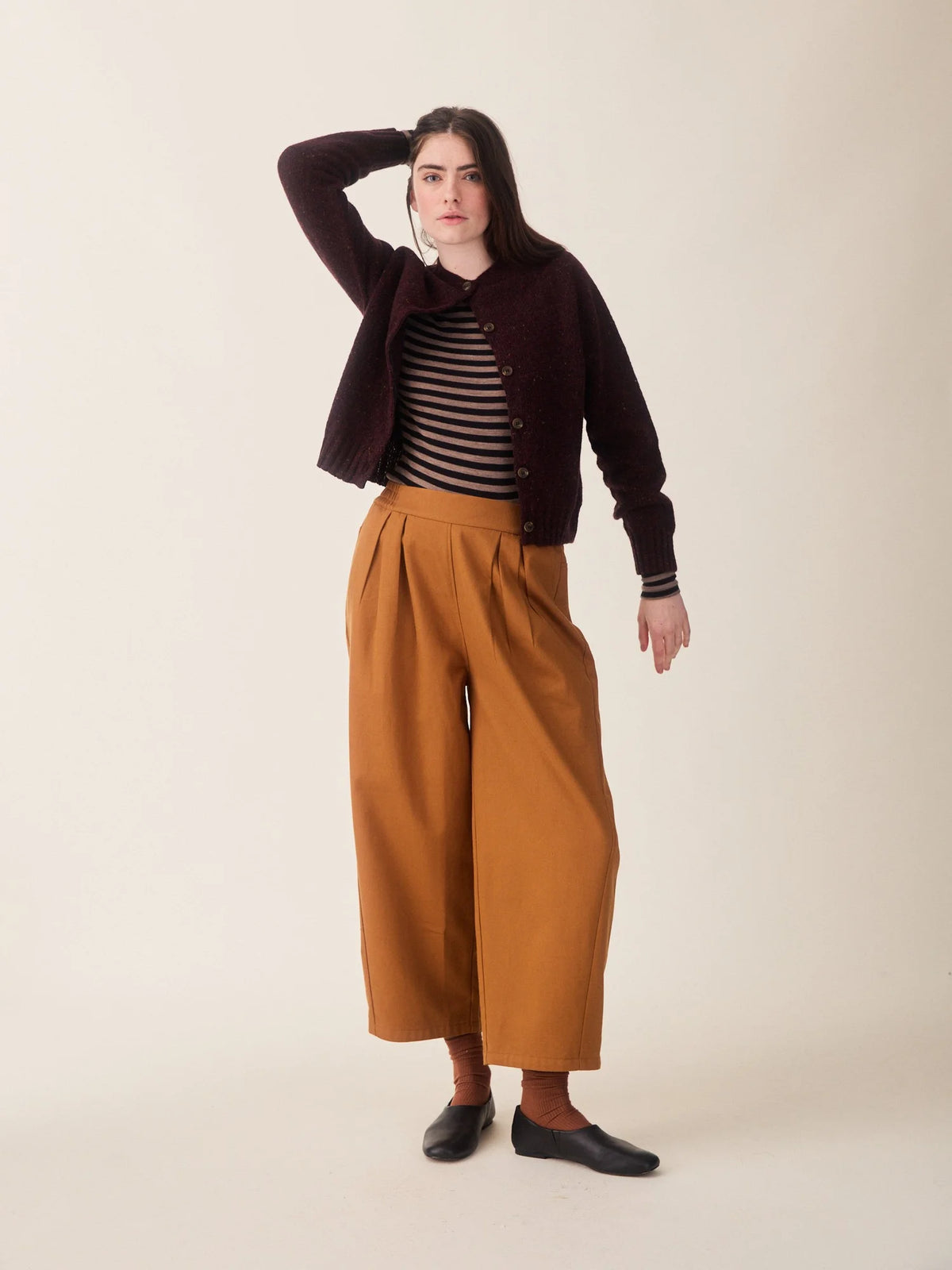 A model stands against a plain beige studio background wearing a dark burgundy knit cardigan over a black and white striped top, paired with wide-leg mustard-brown trousers, brown socks, and black slip-on shoes