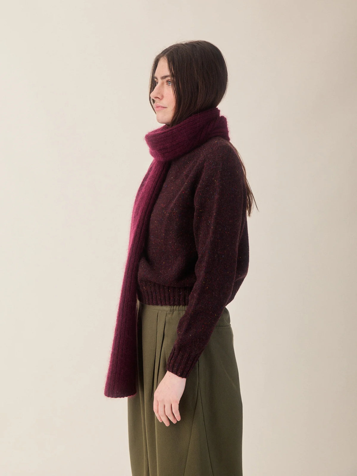 A woman shown in side profile wearing a dark burgundy knit sweater, a matching thick knit scarf, and olive green wide-leg trousers, set against a plain light beige studio background.