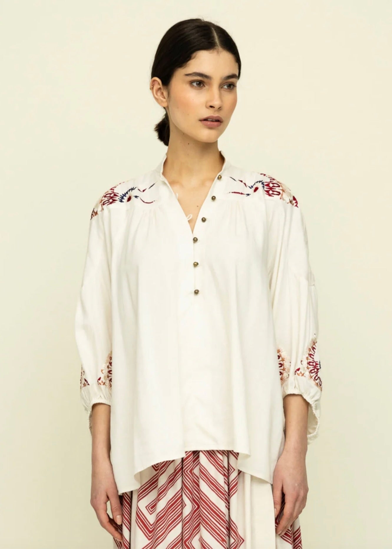 Front view of Side view of woman with dark hair tied back wearing cream colored blouse with colorful embroidery on the shoulders and red and white skirt.