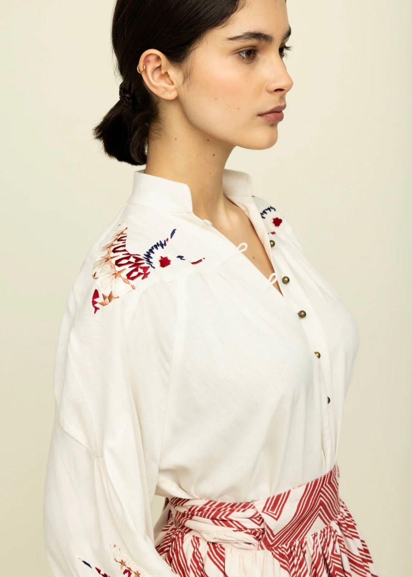 Side view of woman with dark hair tied back wearing cream colored blouse with colorful embroidery on the shoulders and red and white skirt.