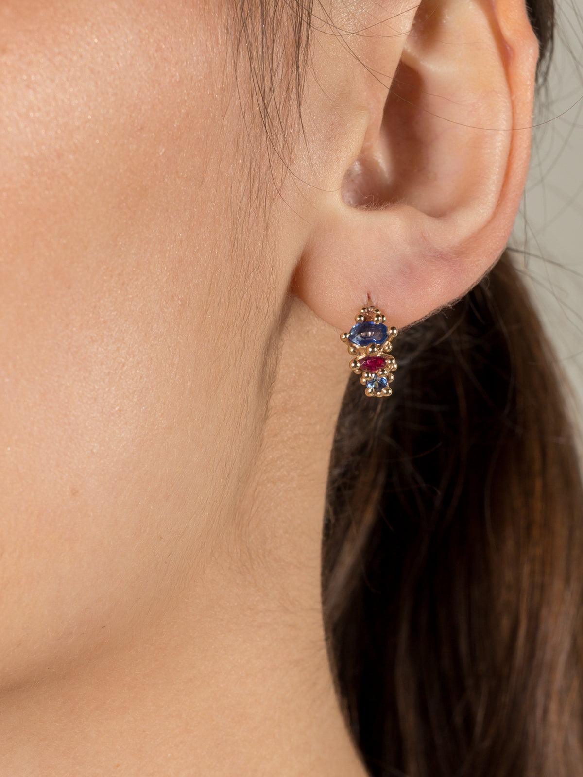 A model shown wearing a yellow gold hoop earring with multi-colored sapphires and rubies in an alternating pattern.