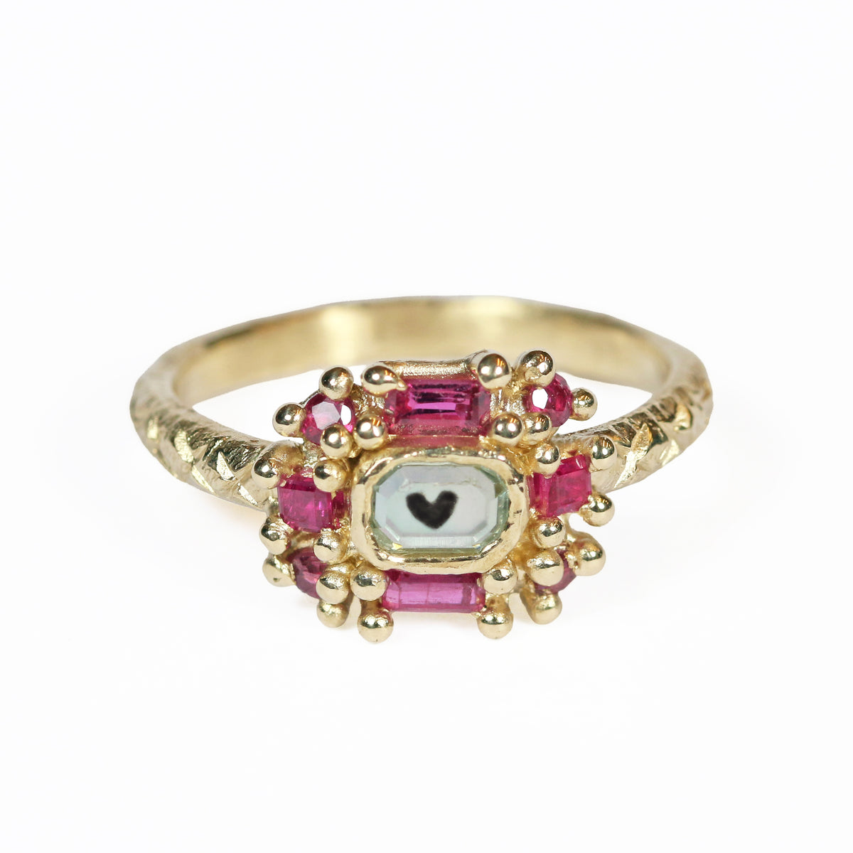 A small heart sits in the center of the yellow gold ring under a white sapphire and surrounded by small rubies.
