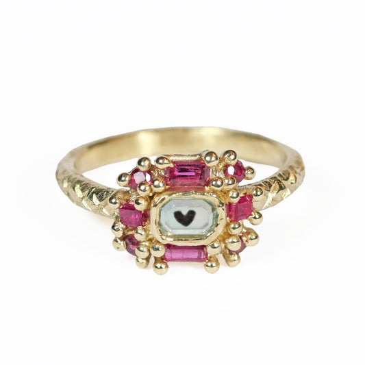 A small heart sits in the center of the yellow gold ring under a white sapphire and surrounded by small rubies.
