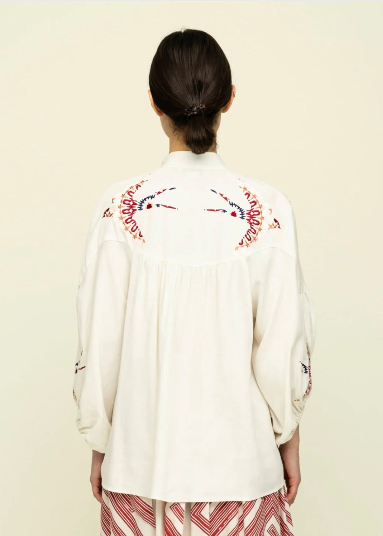 Rear view of Side view of woman with dark hair tied back wearing cream colored blouse with colorful embroidery on the shoulders and red and white skirt.
