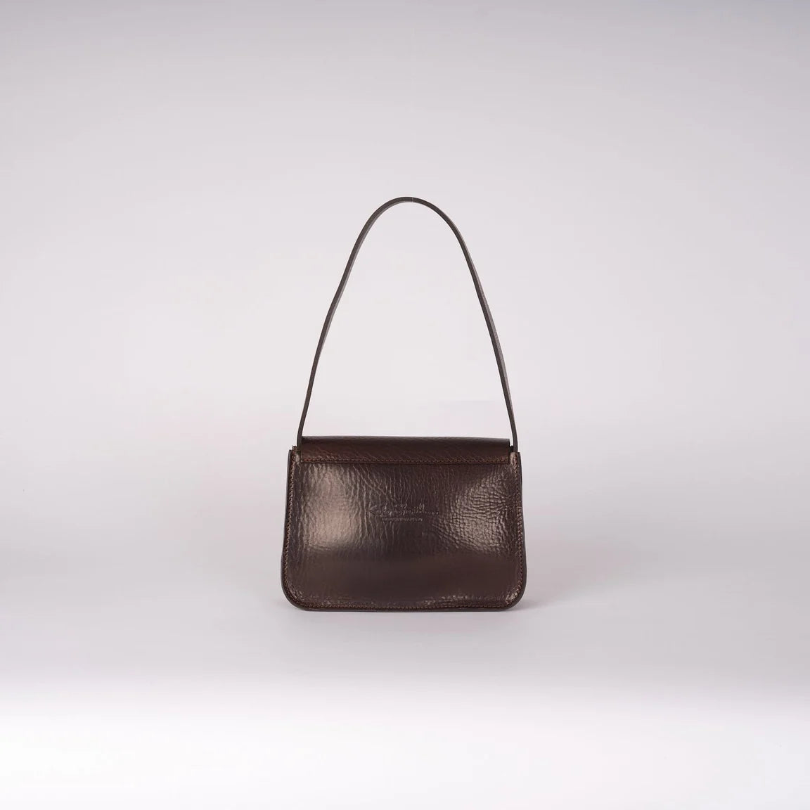 Reverse side view of a dark brown textured leather shoulder bag with a slim matching leather strap, photographed against a neutral light gray studio background.