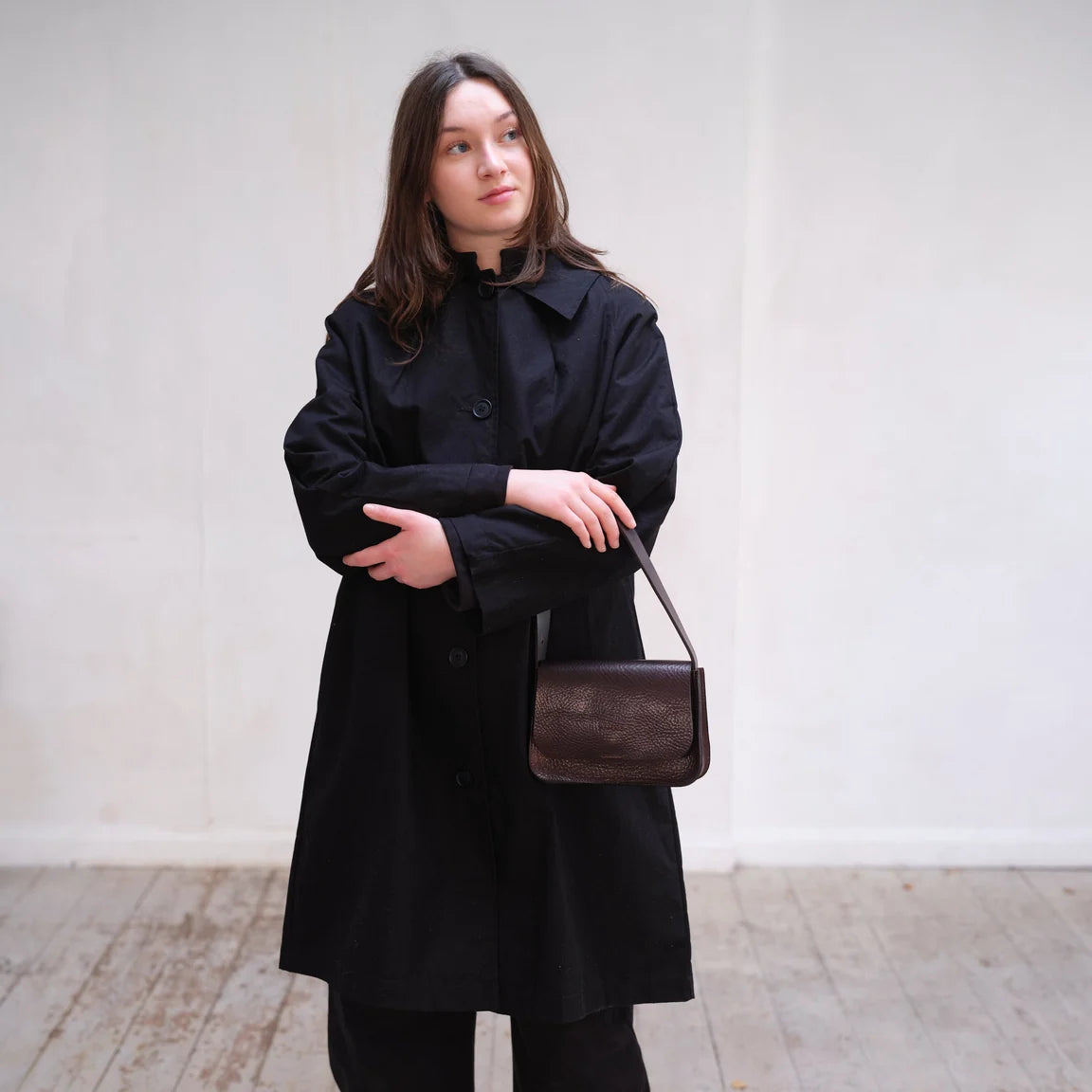 Woman wearing black raincoat with a dark brown textured leather shoulder bag with a slim matching leather strap, photographed against a neutral light gray studio background.