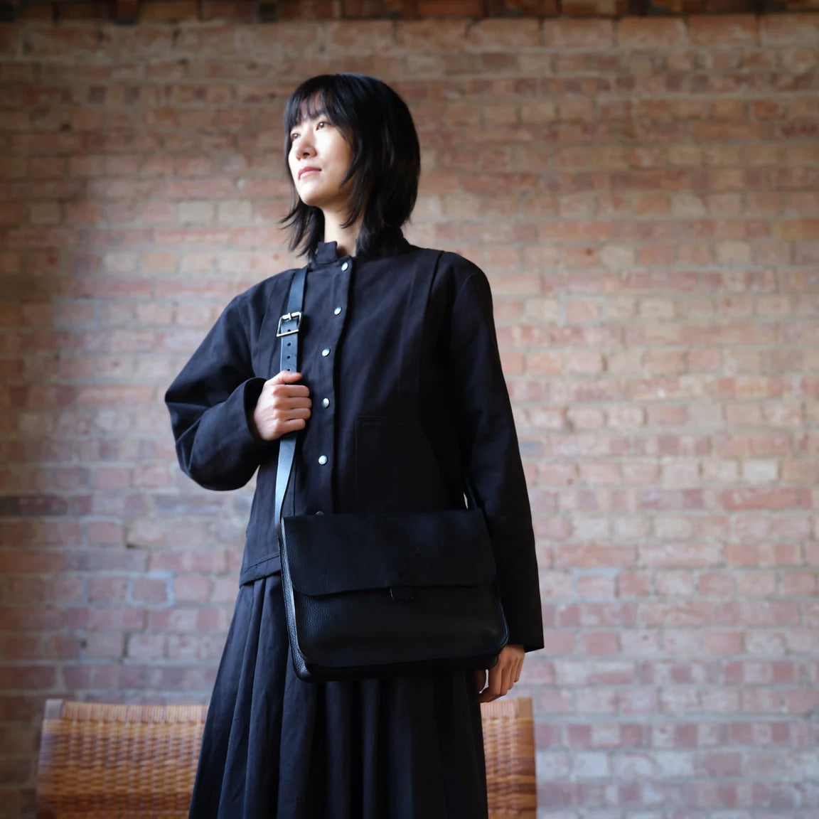 Person dressed in black with black leather shoulder bag standing in front of brick wall